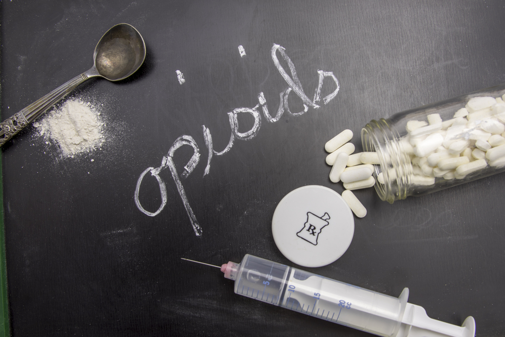 Opioids written in chalk on blackboard with crushed powder, spoon, syring and prescription vial.
