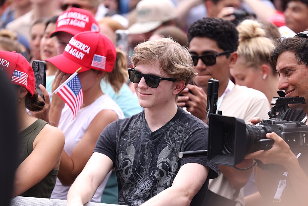 Dean Withers, a young man with blonde hair and sunglasses, sits in a crowd at a political event. He wears a dark patterned shirt while surrounded by red MAGA hats and cameras.
