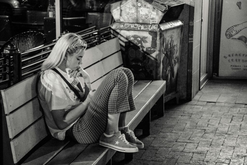 A black and white image of a young woman sitting curled up on a bench, focused on her phone, her body language showing quiet withdrawal.