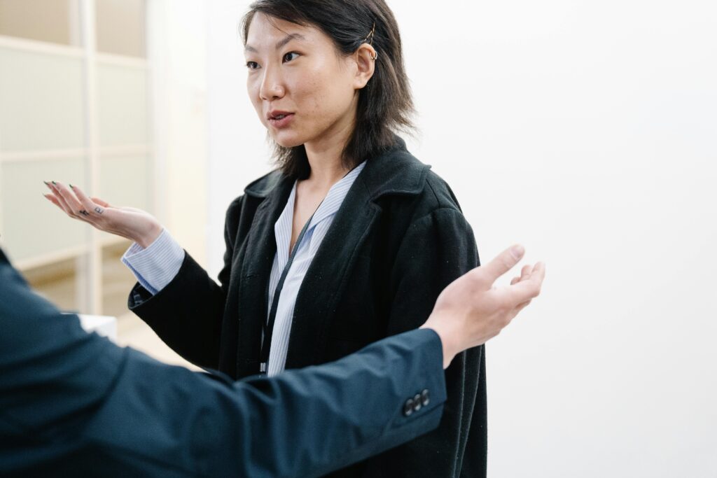 Woman in black blazer gesturing with both hands while speaking to another person in professional setting.