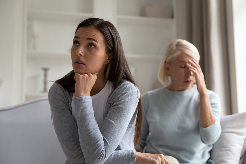 Annoyed angry millennial adult girl sit on couch separate from distressed senior mom thinking of family problems or misunderstanding, pensive grownup daughter having argument with middle-aged mother