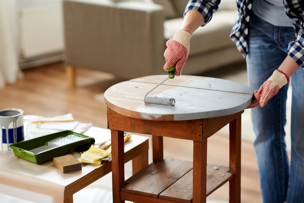furniture renovation, diy and home improvement concept - close up of woman in gloves with paint roller painting old wooden table in grey color