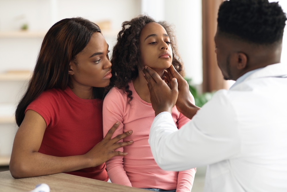 Back view of african american doctor palpating school girl throat at clinic, black mother and sick daughter visiting pediatrician together. Cold and flu, coronavirus, tonsillitis in children concept