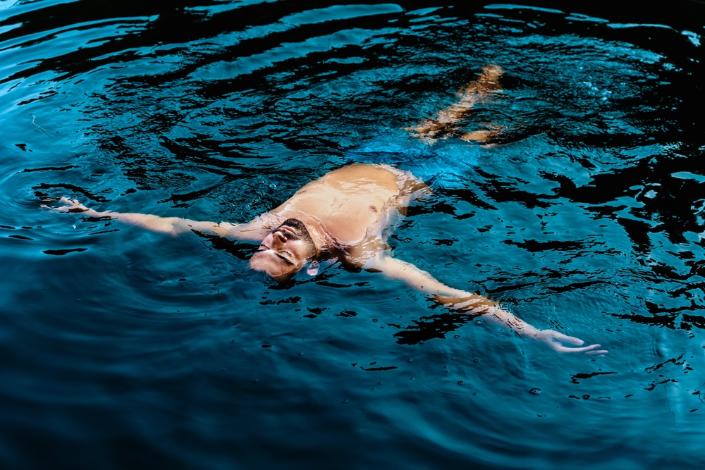 A young bearded man floats calmly on the surface of a dark blue body of water, with his arms outstretched and his eyes closed.