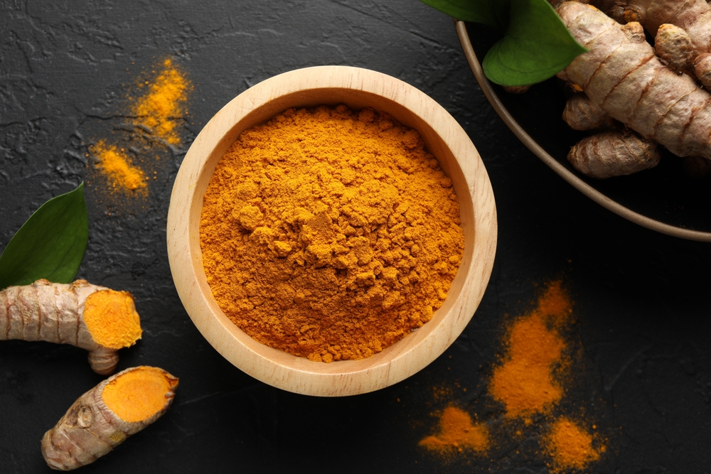 Aromatic turmeric powder in bowl, fresh roots and leaves on black table, flat lay
