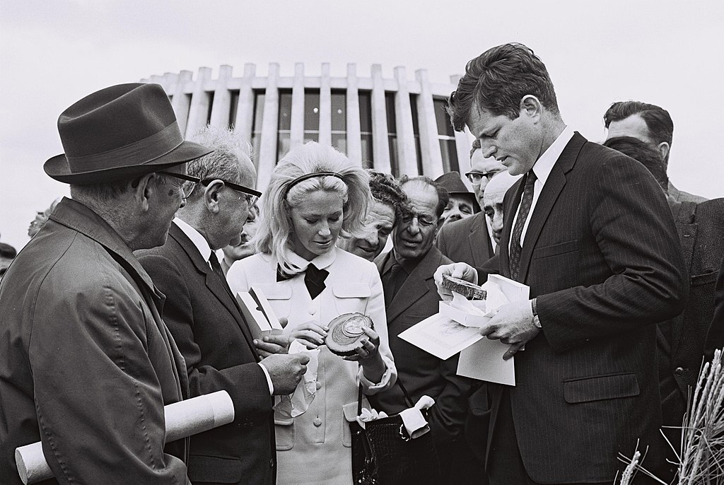 Black and white photo of Joan Kennedy in a light-colored dress with bow collar examining a commemorative medallion while standing beside Ted Kennedy in a pinstriped suit holding papers. Ya'acov Tzur, Chairman of the Jewish National Fund, and other officials in suits and hats stand to the left. 