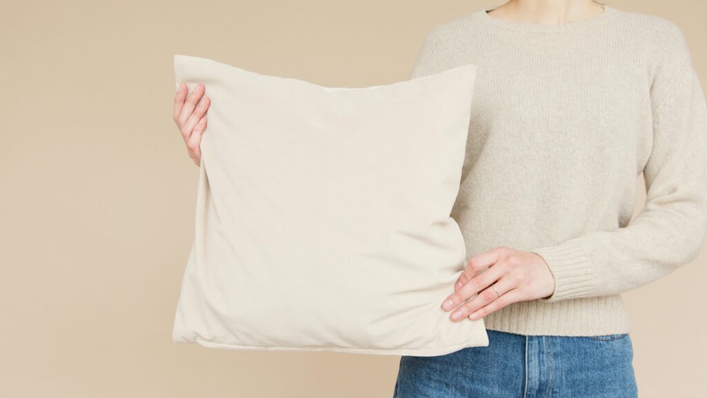 Person holding white pillow in cream sweater, against a cream background.
