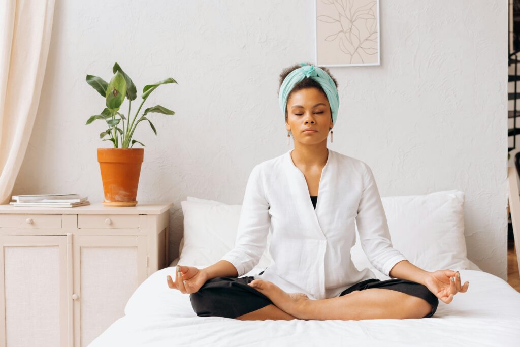 Woman in lotus position on bed wearing white robe and turquoise headwrap with potted plant on nightstand beside her.