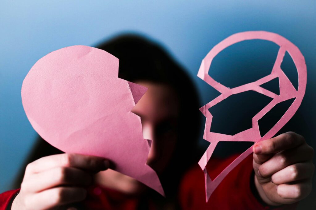 Person holding pink paper heart and geometric face cutout in front of their own face against blue background.