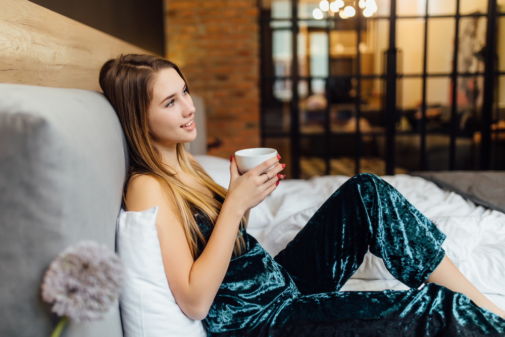 Morning coffee is my daily routine, cute woman with cup of coffe lying on bed at modern luxury apartment.