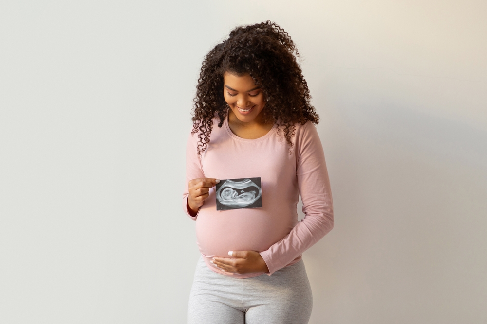 Maternity Concept. Happy Black Pregnant Woman Holding Ultrasound Photo Of Her Baby And Tenderly Embracing Belly While Standing Against White Wall At Home, Expectant Mother Keeping Sonography Scan