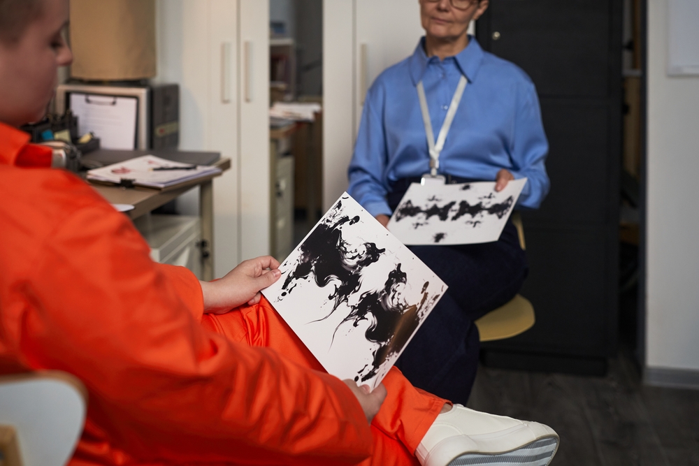 Young adult Caucasian woman wearing orange prison uniform, sitting in office holding inkblot test card while middle aged Caucasian woman psychologist observing during prison therapy session