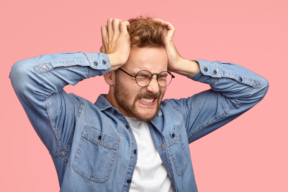 Young unshaven stressful man suffers from headache, keeps hands on temples, clenches teeth, has terrible pain, closes eyes, wears round spectacles, dressed in denim shirt, isolated over pink wall
