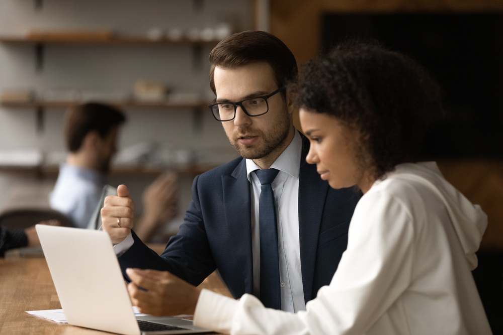 Serious millennial businessman manager expert explain african woman client project details using laptop. Confident young male mentor teach black female colleague instruct student intern at workplace