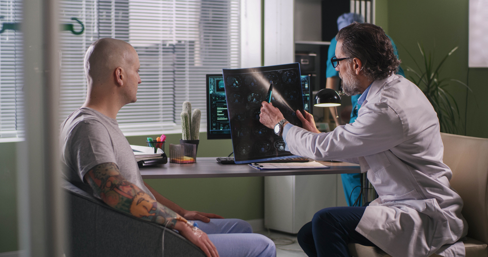 Mature oncologist explaining condition of incurable brain cancer to bald patient while sitting near computer during chemotherapy