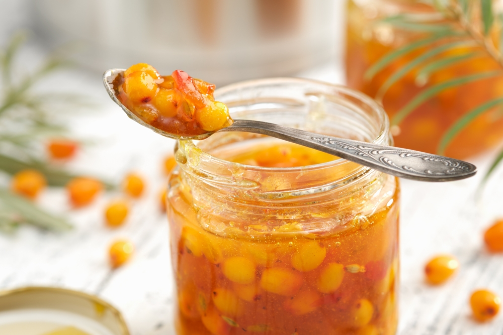 Jar of healthy sea buckthorn jam. A spoonful of buckthorn jam close up. A pot, jam jar and sea buckthorn berries in the background.