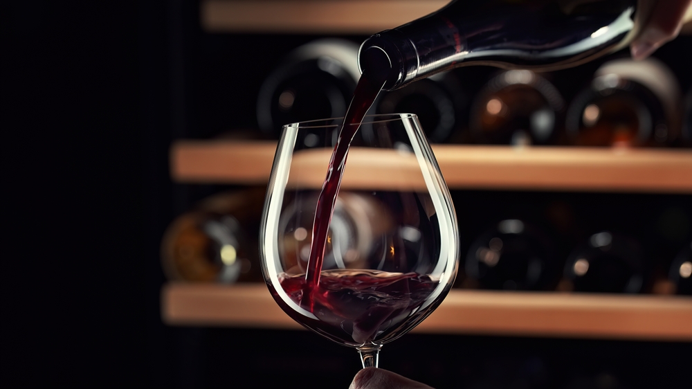 Close up female hand pouring red wine to glass. Wine expert, sommelier tasting, rating and drinking wine, bottles in background. Restaurant, wine bar, waitress.