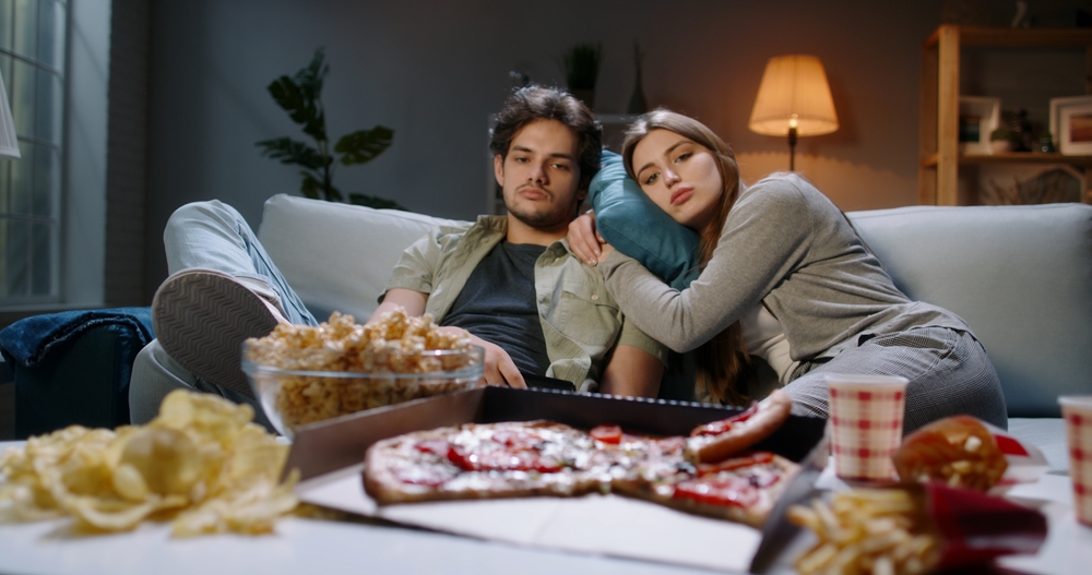 Young caucasian couple extremely frustrated. Bored students are sitting in front of tv, switching channels with remote and eating unhealthy food, tired after hard day
