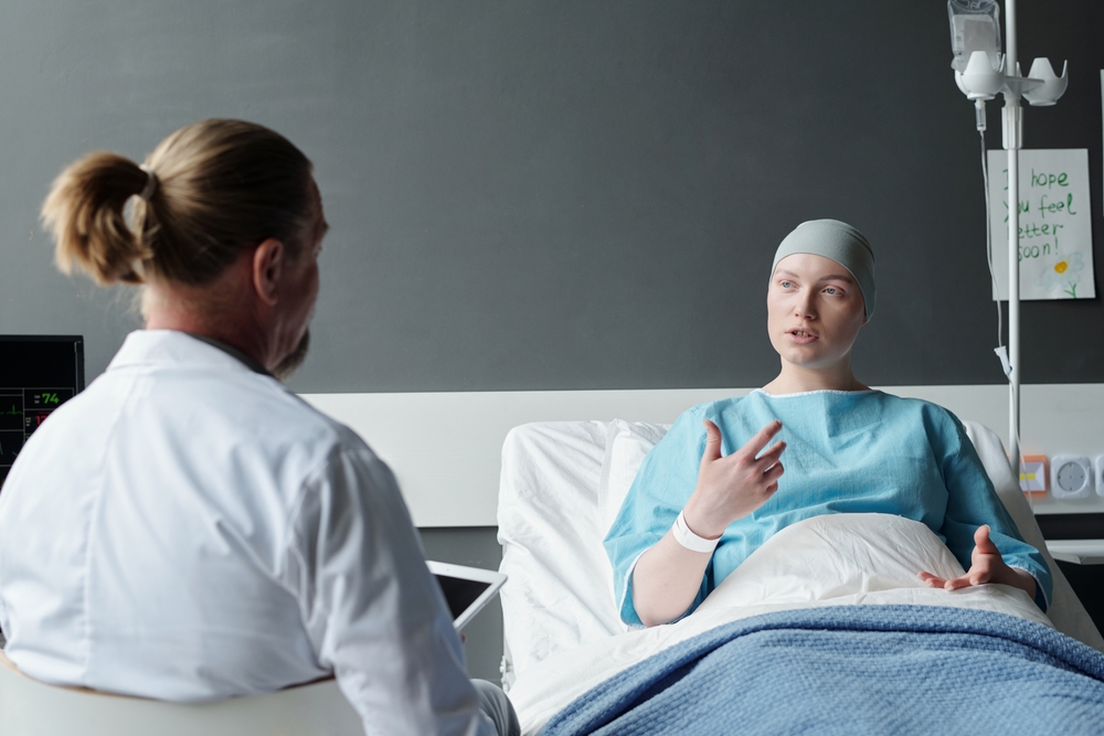 Young female patient explaining experienced oncologist her symptoms while sitting in bed and looking at male doctor in lab coat in clinics