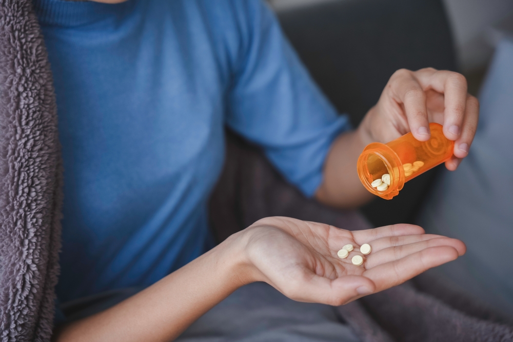 Sick woman is sitting on a couch under a blanket and pouring pills from a prescription bottle into her hand