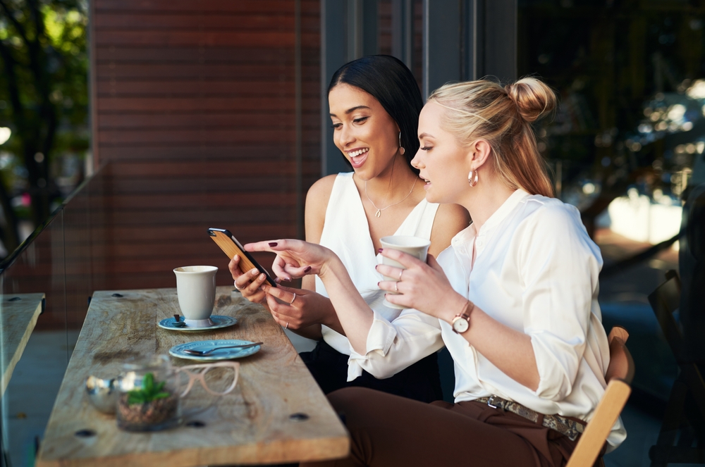 Pointing, cellphone and friends in cafe, smile and choice for women, gossip and happy to relax in weekend. Customer, enjoy and girls with beverage, conversation and break in morning and students
