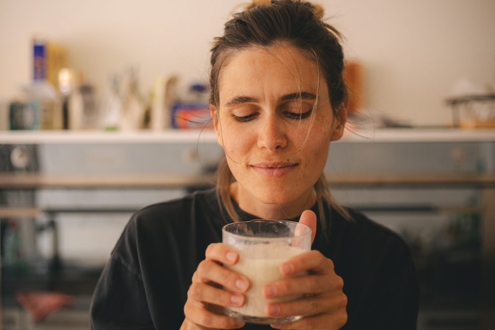 Woman standing on the kitchen, holding glass of warm milk drink and enjoy of flavor, smell it.
