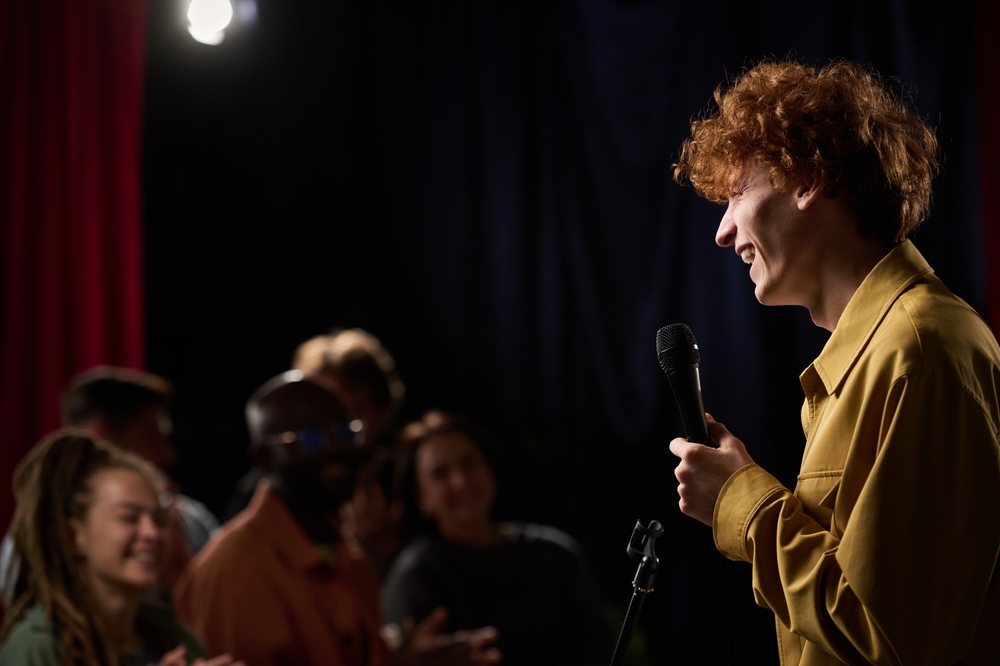 Person with curly hair holding microphone while performing stand-up comedy on stage. Audience consisting of diverse people enjoying show and laughing together in dark room