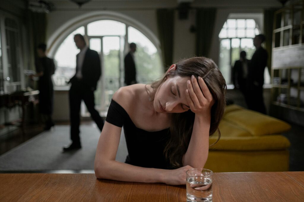 A young woman sits at a table with her head resting in her hand, eyes closed in exhaustion. A small glass sits in front of her while men in suits converse in the background near an arched window.