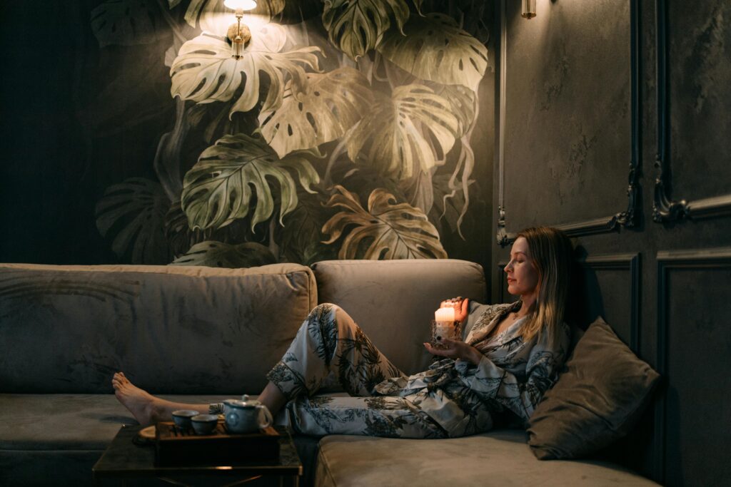 A woman in patterned pajamas sits on a velvet couch holding a lit candle. Tropical leaf wallpaper and a tea tray create a cozy atmosphere.