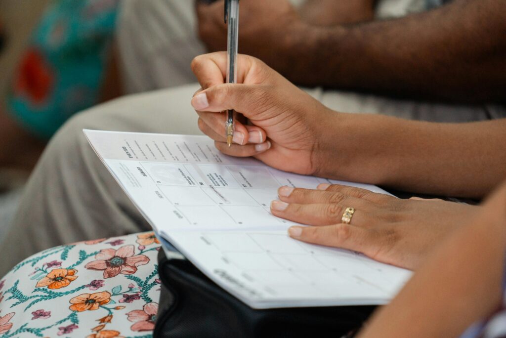 Close-up of hands writing in a planner with a pen. The person wears a floral print dress and a gold ring.