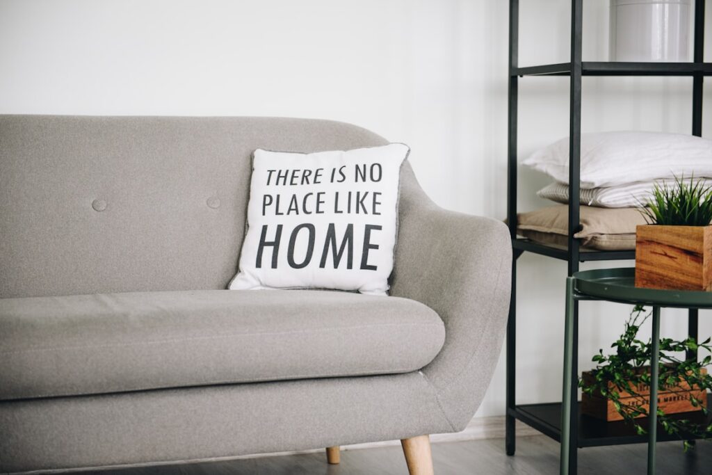 A gray couch with a white pillow reading "There Is No Place Like Home." A shelving unit with plants and linens stands beside it.
