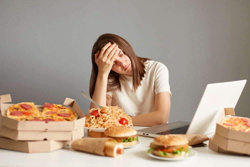 Portrait of tire overworked woman working on laptop computer, sitting surrounded with big variety of fast food, keeps palm on forehead, isolated over gray background.