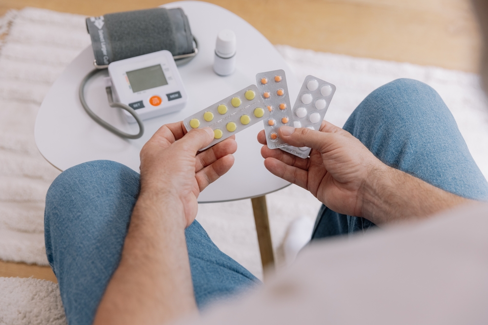 Close-up of elderly man sitting sofa at living room measures blood pressure with device. Senior male holding pills and drops drugs. Health and self-care concept, copy space