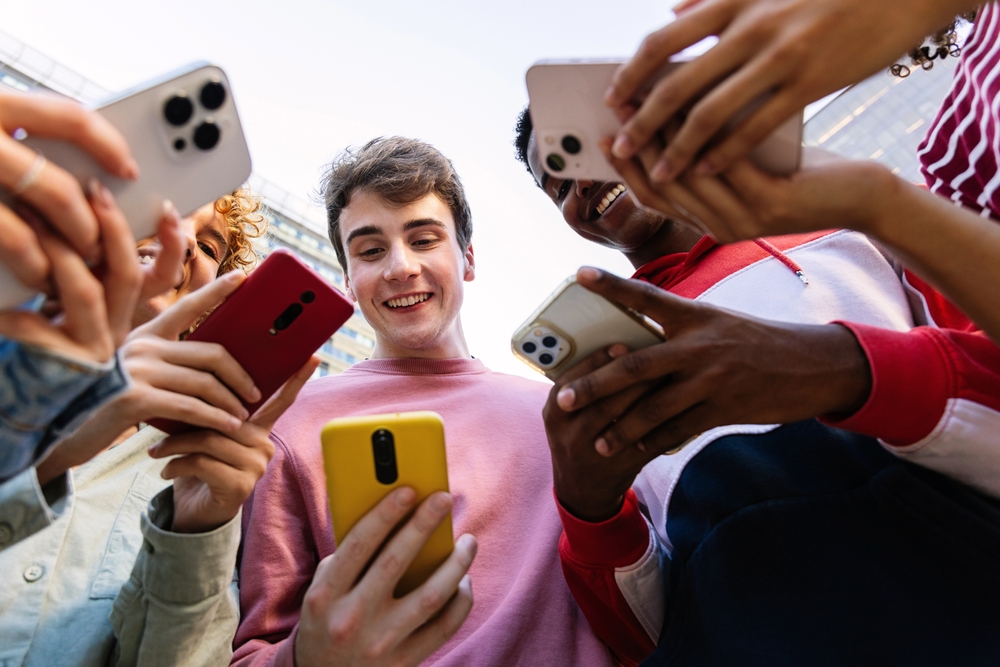 Young group of teen friends using mobile phone device standing in circle outdoors. Gen z students addicted to social media app, betting or playing video game on platform online.