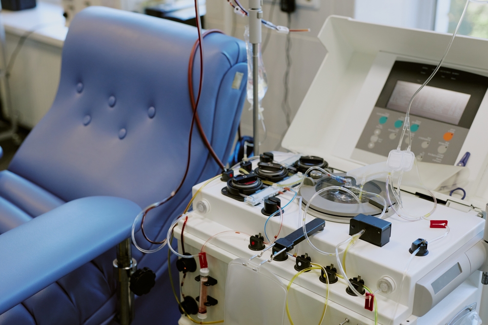 Medical devices arranged for hemodialysis in hospital room. Blue cushioned chair prepared for patient, with advanced dialysis machine beside it having tubes and controls visible