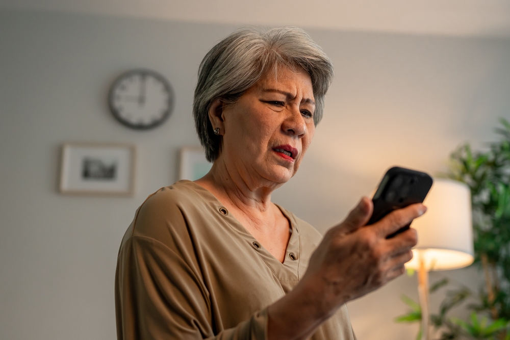 Depressed Asian senior woman sit alone on bed and looking at smartphone. Elderly older mature grandmother feel lonely and sad, missing family and relative while spend time in house after retirement.