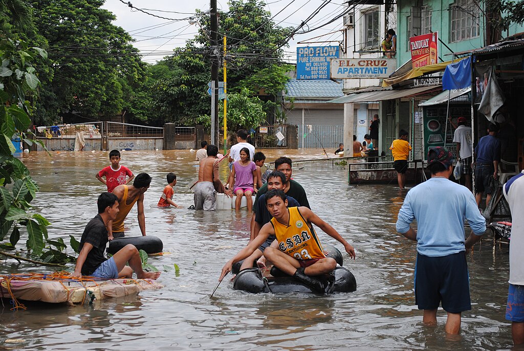Residents wading and floating through chest-high floodwater in a residential neighborhood, using inner tubes and improvised rafts to navigate between partially submerged buildings.