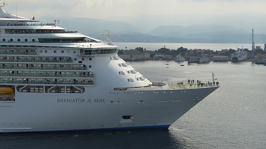 The bow of Royal Caribbean's Navigator of the Seas enters a harbor, with passengers standing on the forward observation deck. The ship's name appears in dark lettering against the white hull, and a coastal town with mountains rises in the misty background.