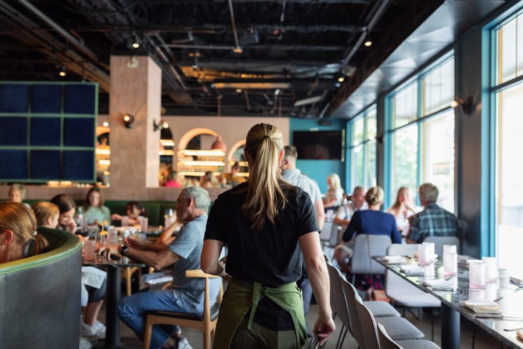 A server with a blonde ponytail walks through a busy restaurant dining room, seen from behind, with guests seated at tables along large windows.
