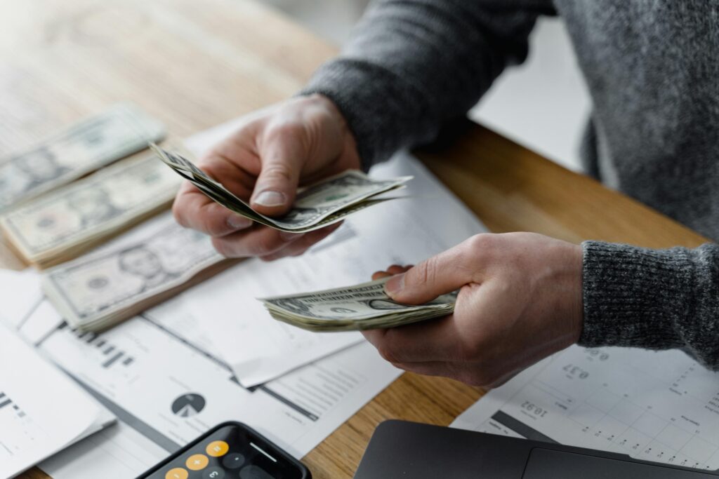 A pair of hands counts a stack of hundred-dollar bills over a desk covered with paperwork, a calculator, and additional bundles of cash.