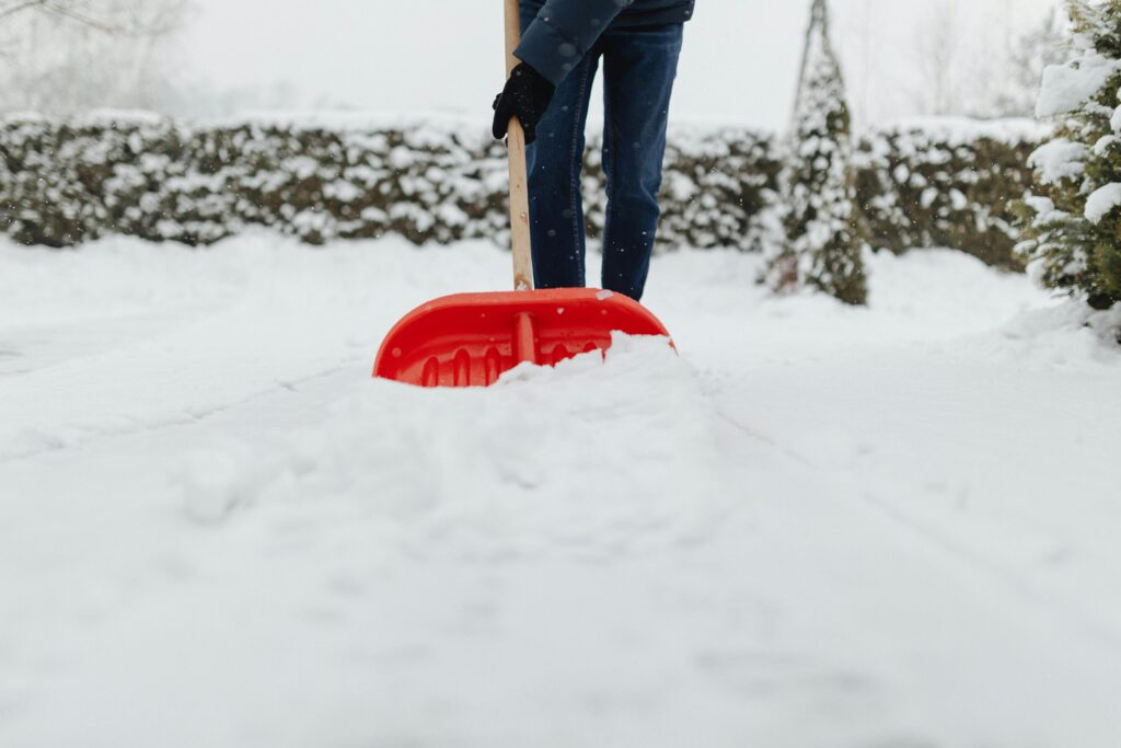 A person in jeans and dark boots pushes a red snow shovel through a thick layer of snow in a backyard, with snow-covered hedges behind them.