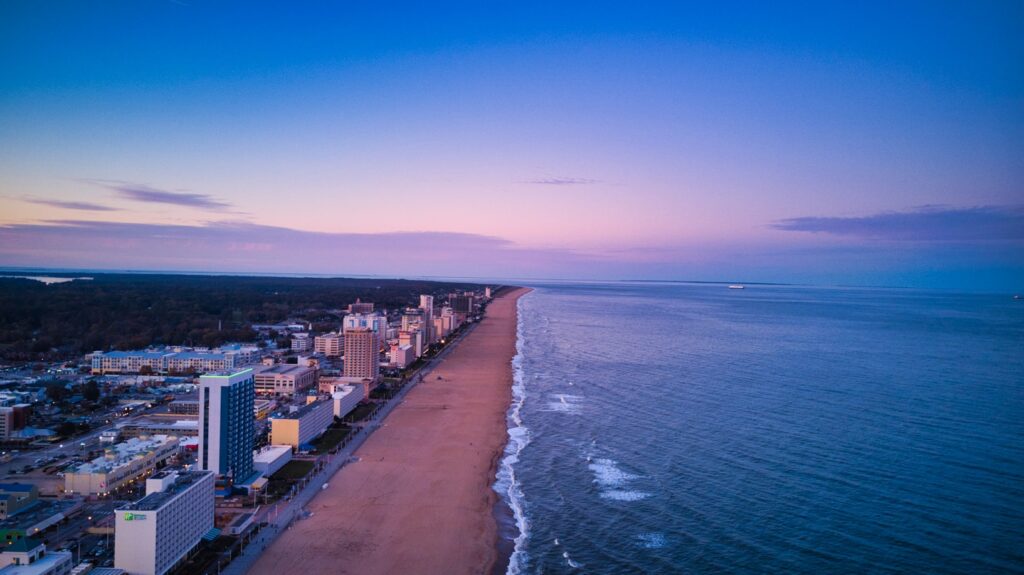 An aerial view of Virginia Beach at sunset, with high-rise hotels lining the shore and gentle waves meeting the sand under a pink and blue sky.