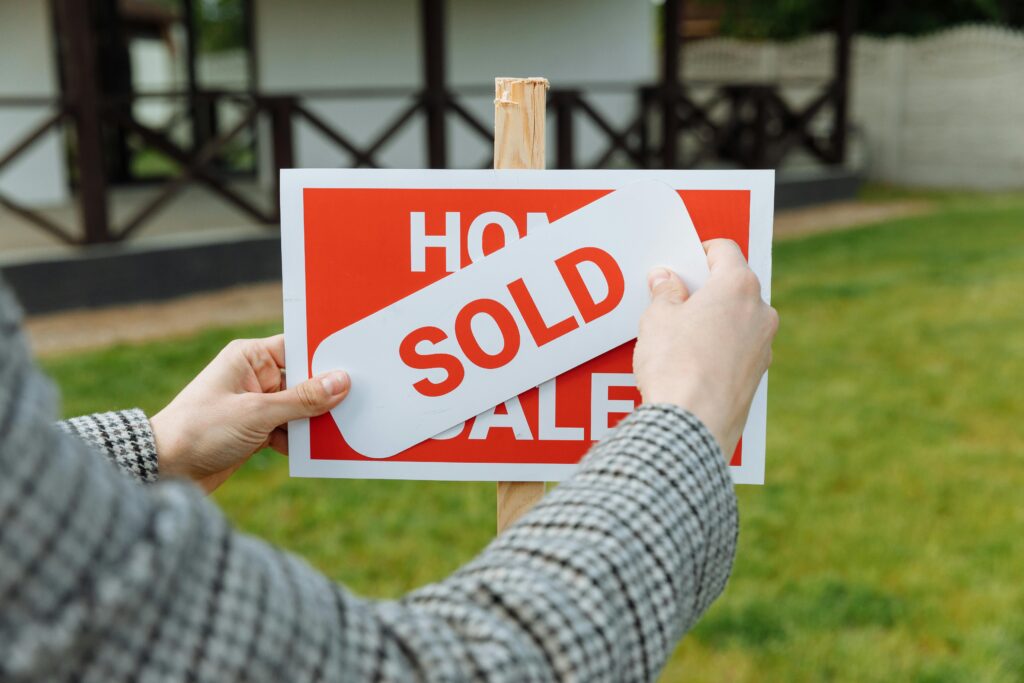 A person in a plaid jacket places a white "SOLD" sticker diagonally across a red "Home for Sale" sign on a front lawn.