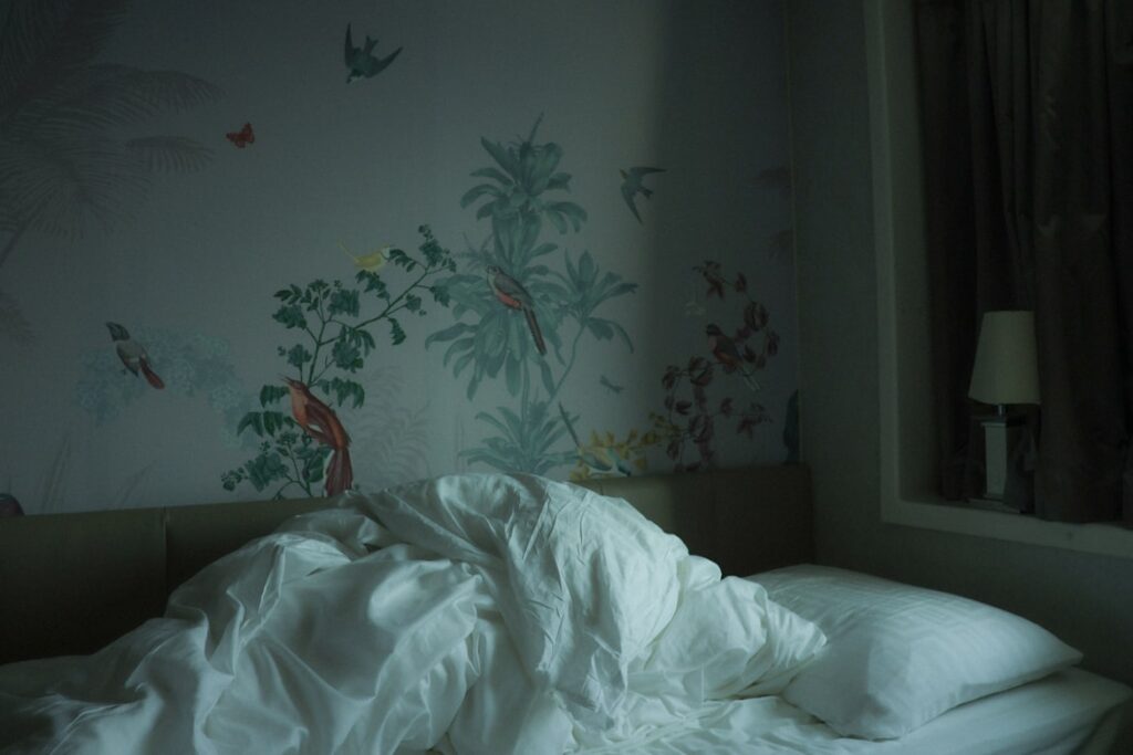 An unmade bed with crumpled white sheets in a dim room, the wall behind it covered in tropical wallpaper with painted birds and palm fronds.