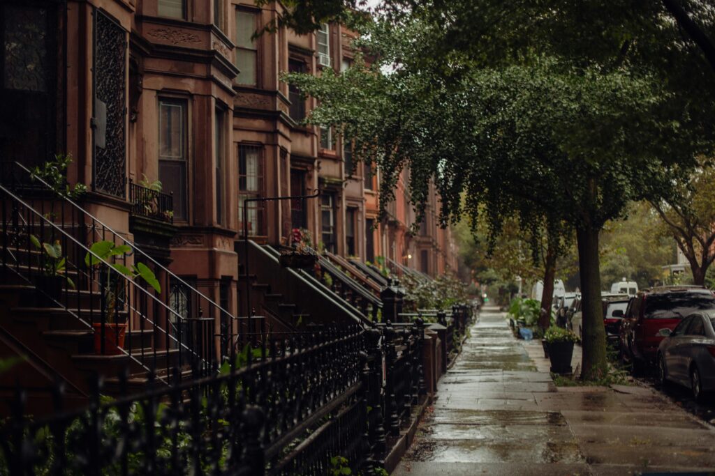 A row of brownstone townhouses on a rainy tree-lined street, with iron railings leading up stone stoops and parked cars along the curb.