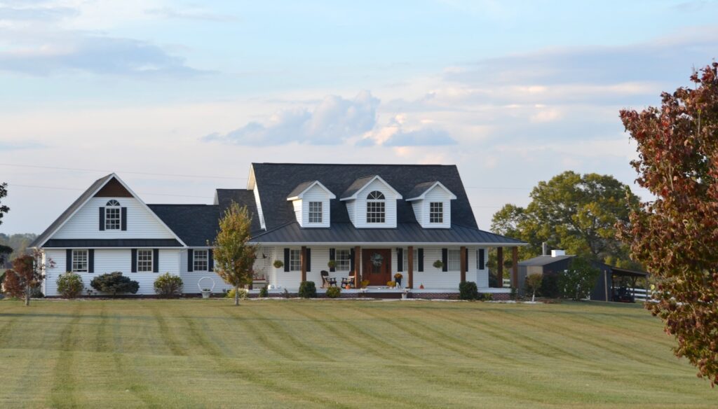 A white farmhouse with black shutters and a wraparound porch, sitting on a large green lawn with a few trees and open sky behind it.