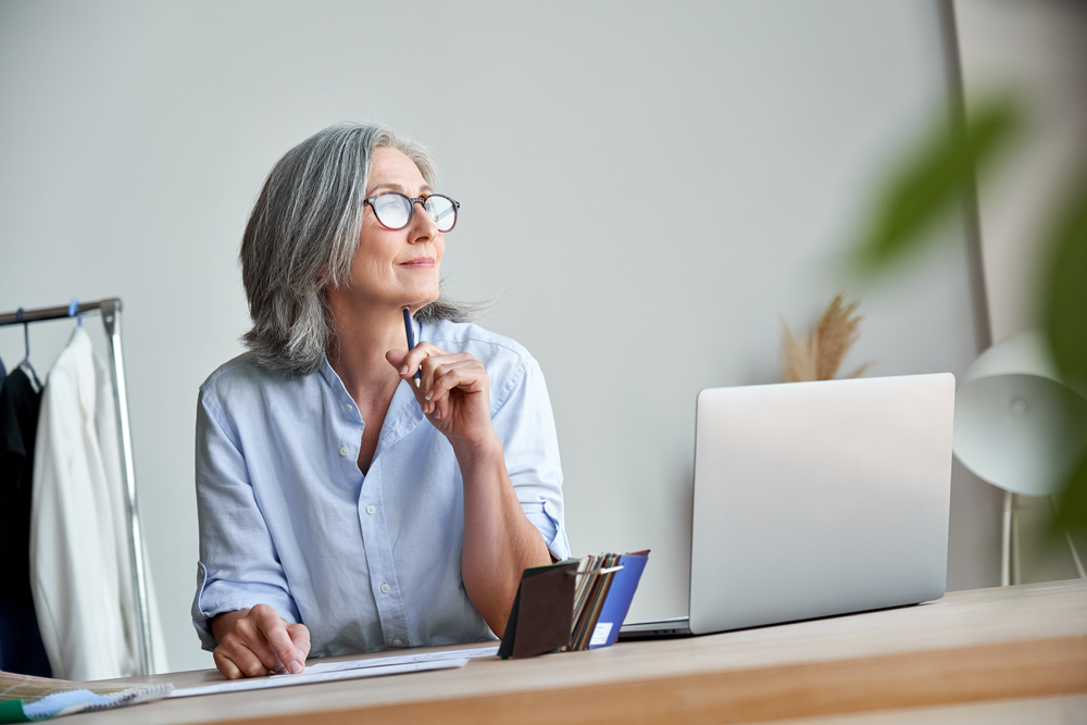 Inspired mature grey-haired woman fashion designer thinking on new creative ideas at workplace. Smiling beautiful elegant classy middle aged older lady small business owner dreaming in atelier studio.