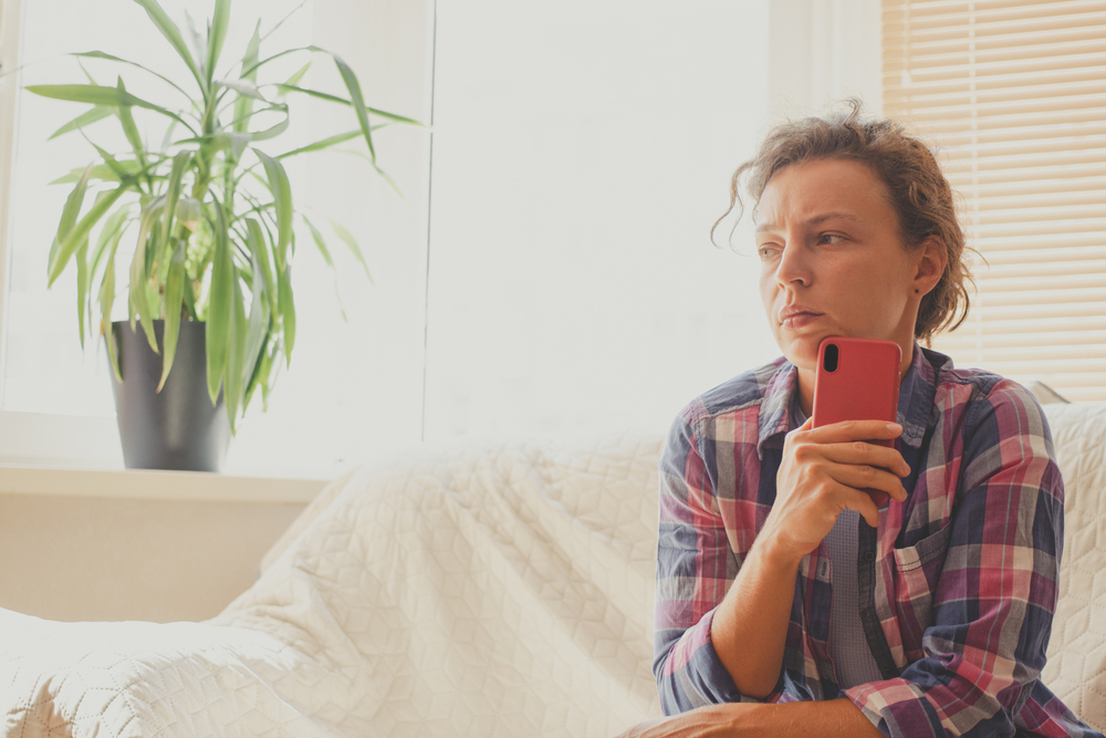 Sad worried young woman feels desperate waiting for call, holding mobile phone sitting on sofa at home, looking to distance. Thinking about personal troubles, break up, heartbreak, treason.Copy space