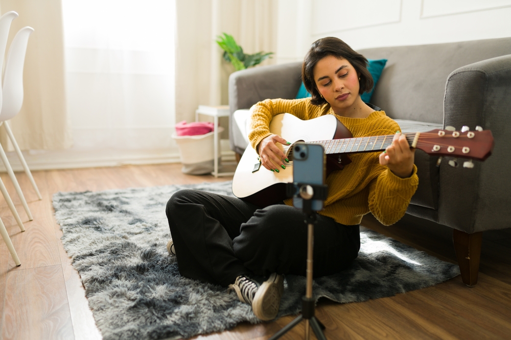 Creative musician practicing and recording music lessons at home, using a smartphone to capture her acoustic guitar performances