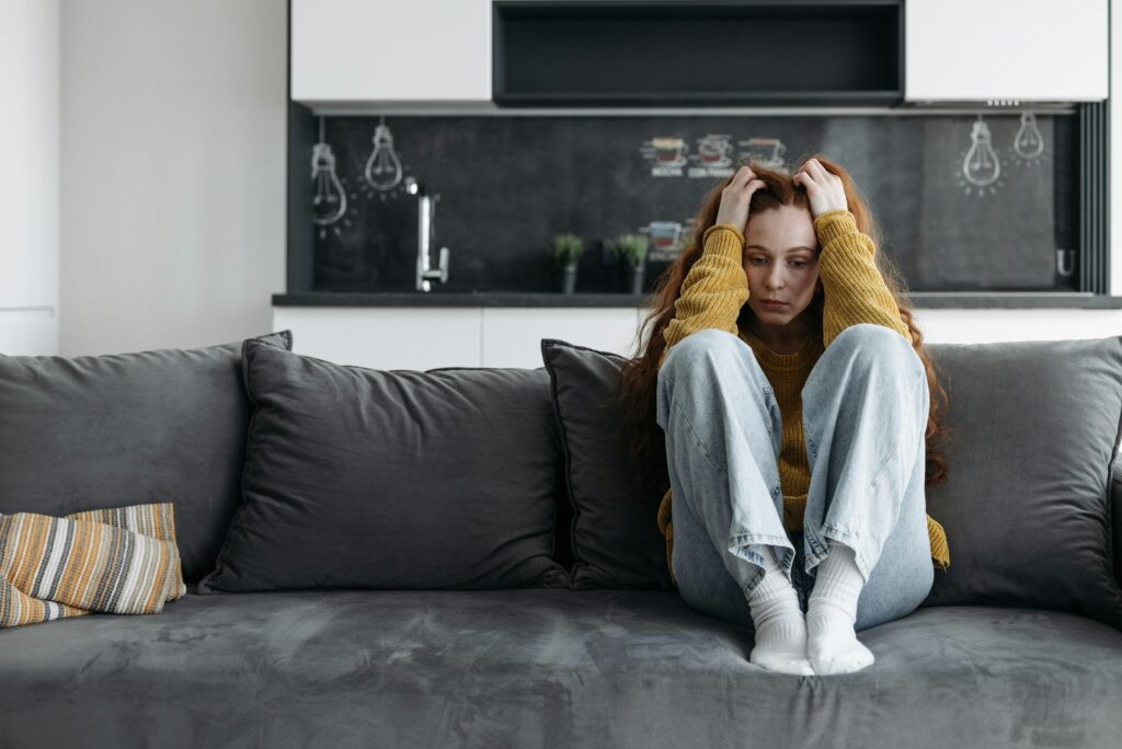 A young red-haired woman sits curled up alone on a grey couch in a modern apartment, both hands gripping her hair, head bowed. She's wearing a yellow sweater and light jeans.